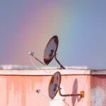 Two rusty satellite dishes on a rooftop against a vibrant rainbow in Tunisia.